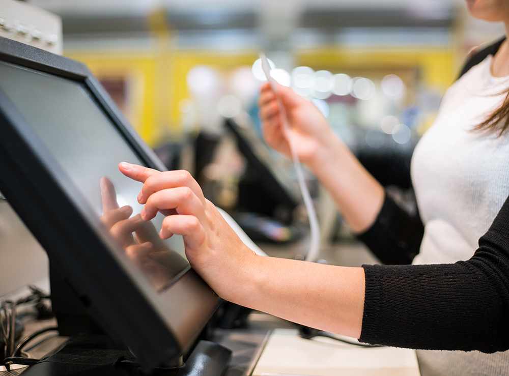 Nahaufnahme einer Person, die eine moderne Touch-POS-Kasse bedient. Der Fokus liegt auf den Händen, eine Hand tippt auf den Bildschirm.
