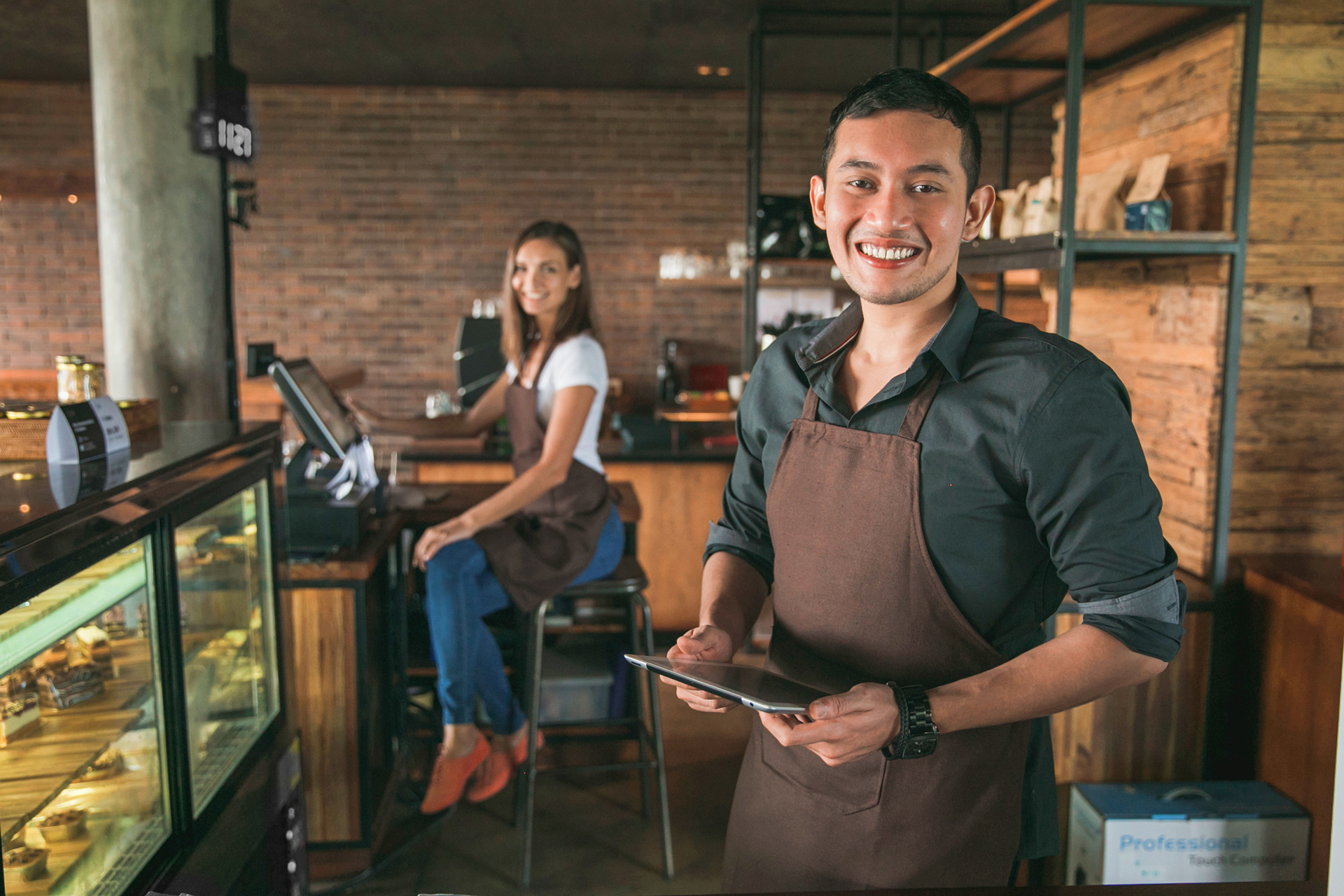 Cafébesitzer nutzt in seinem Coffeeshop ein Tablet, während eine Kellnerin im Hintergrund sitzt.