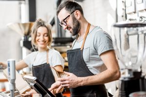 Zwei Verkäuferinnen in Uniform füllen Tüten mit Kaffeebohnen in einem Kaffeegeschäft. Sie stehen vor der Kasse.